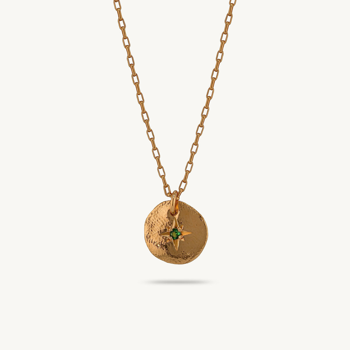 A gold chain necklace featuring a textured circular pendant with a small emerald central stone, shown against a light cream background.