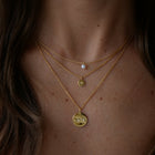 A close-up of a woman's neck wearing three layered gold necklaces: a delicate chain with a single white pearl, a chain with a small gold disc charm, and a longer chain with a larger gold disc pendant featuring a starry sky design.