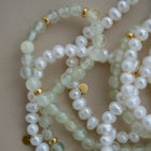 A close-up of a beaded strand featuring pale green translucent gemstone beads, white pearl-like round beads, and small gold spacer beads, arranged on a neutral beige background.