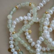 A close-up of a beaded strand featuring pale green translucent gemstone beads, white pearl-like round beads, and small gold spacer beads, arranged on a neutral beige background.