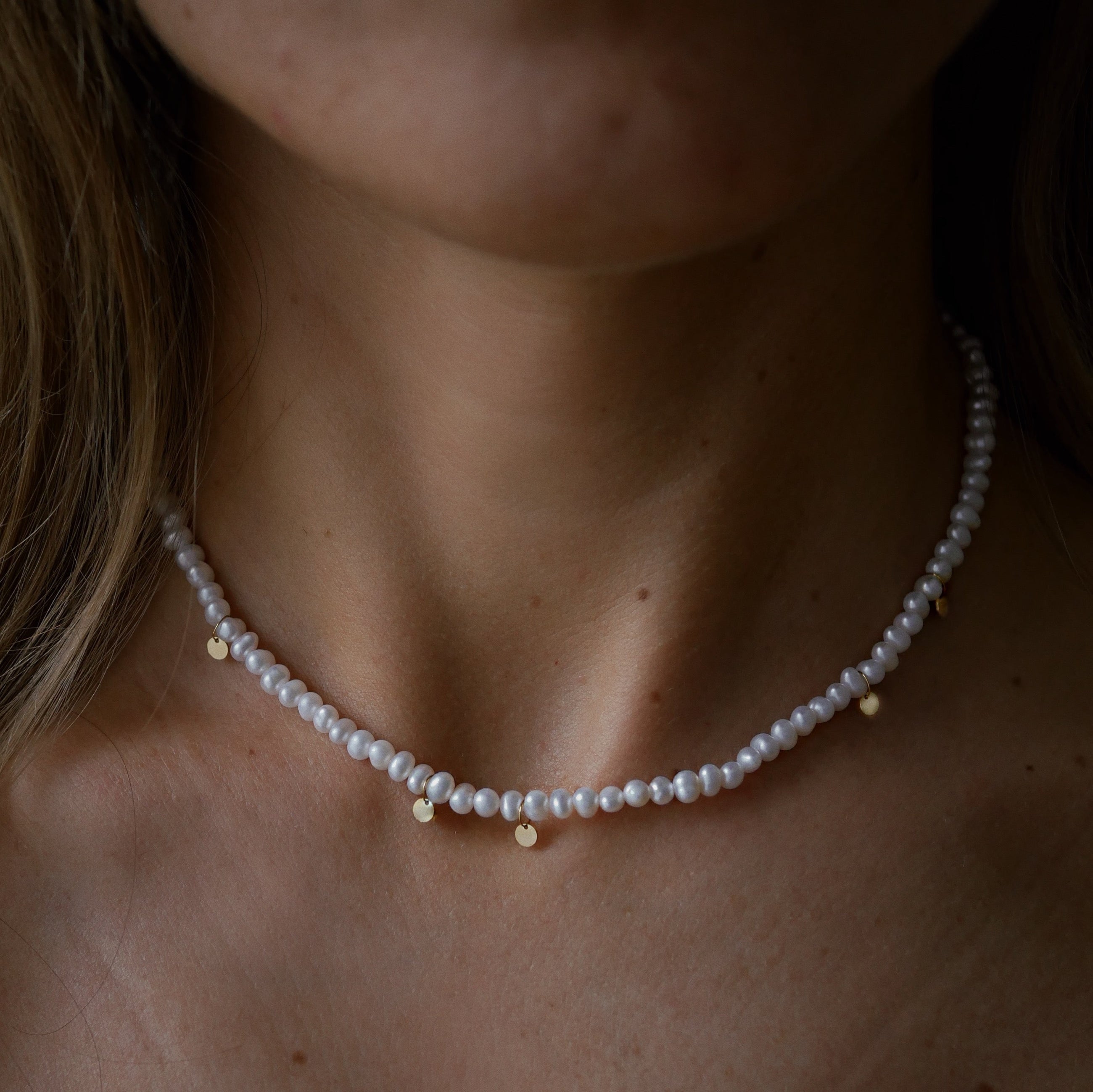 A close-up of a model's neck and collarbone wearing a delicate white pearl necklace with small gold-plated silver accents.