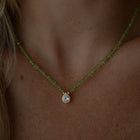A close-up of a person's neck and chest wearing a delicate necklace featuring a strand of small, faceted peridot beads with a gold-tone setting holding a teardrop-shaped zirconium.