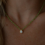 A close-up of a person's neck and chest wearing a delicate necklace featuring a strand of small, faceted peridot beads with a gold-tone setting holding a teardrop-shaped zirconium.