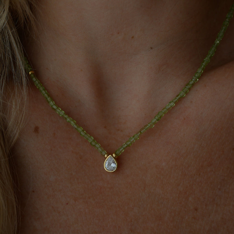 A close-up of a person's neck and chest wearing a delicate necklace featuring a strand of small, faceted peridot beads with a gold-tone setting holding a teardrop-shaped zirconium.