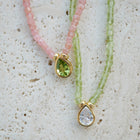 Two beaded necklaces with gold teardrop pendants, one in pink beads with a green gemstone and one in light green beads with a clear gemstone, displayed on a textured white surface.