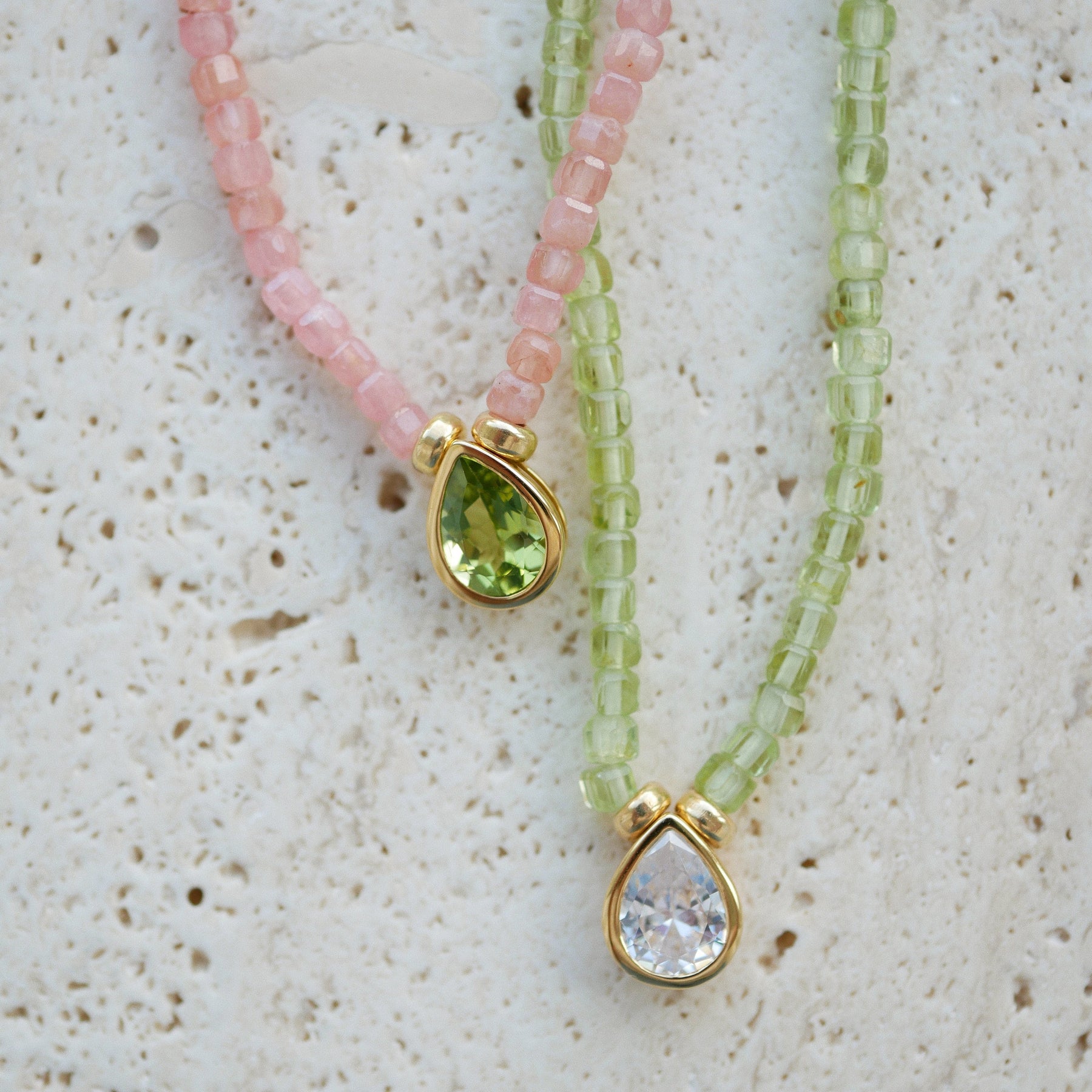 Two beaded necklaces with gold teardrop pendants, one in pink beads with a green gemstone and one in light green beads with a clear gemstone, displayed on a textured white surface.