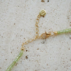 Close-up of a delicate gold chain necklace extender with a lobster clasp, a round charm, and light green faceted beads on a textured beige stone surface.