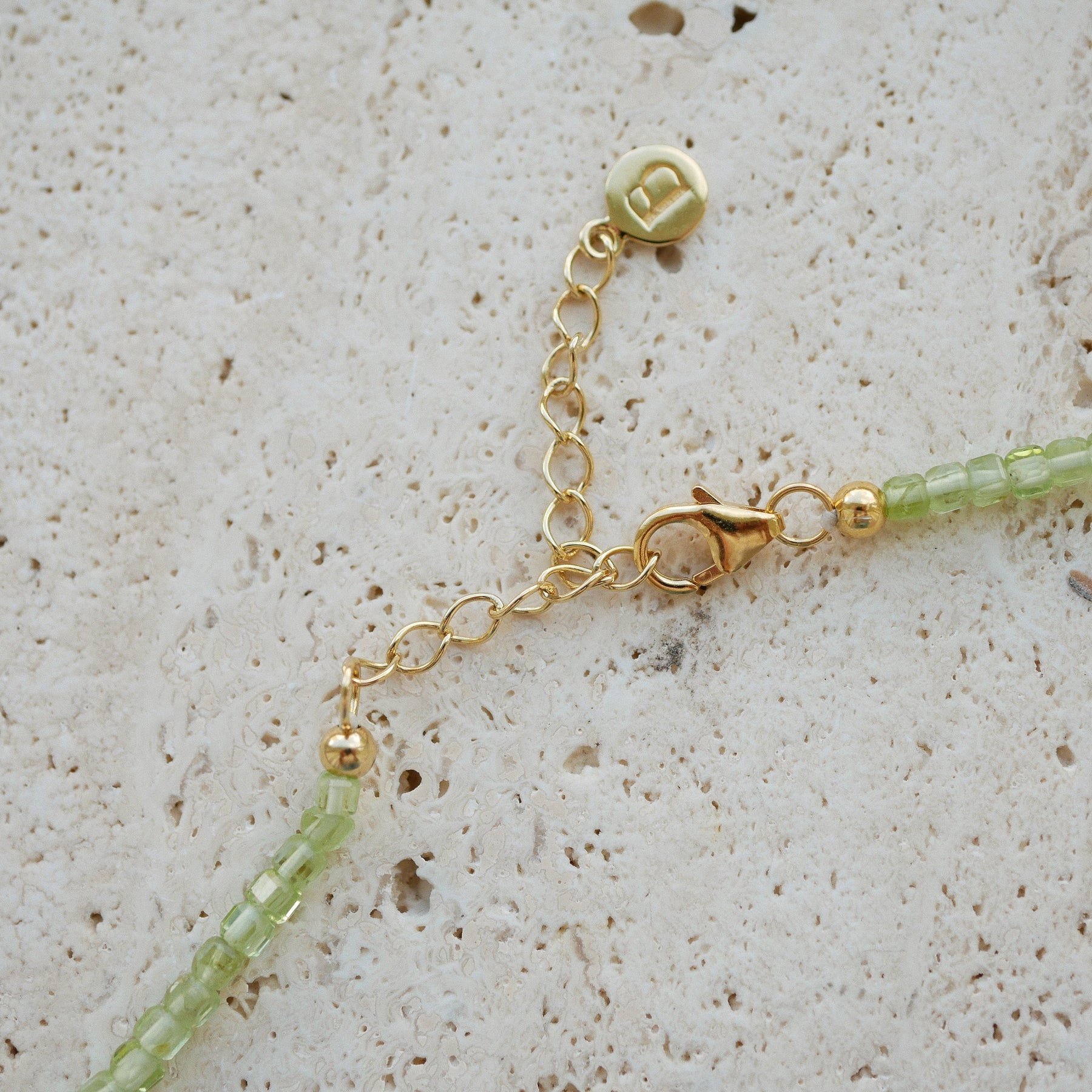 Close-up of a delicate gold chain necklace extender with a lobster clasp, a round charm, and light green faceted beads on a textured beige stone surface.