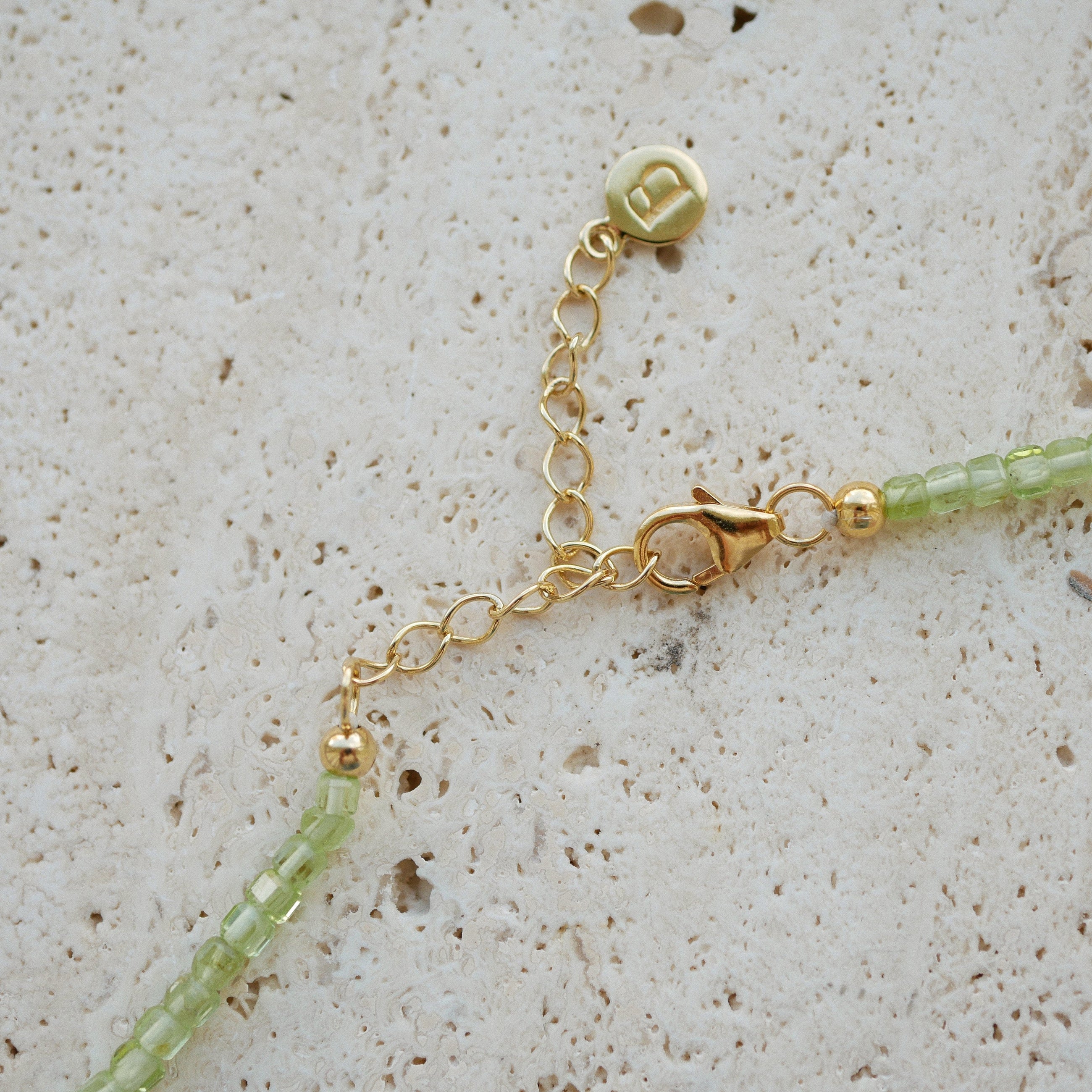 Close-up of a delicate gold chain necklace extender with a lobster clasp, a round charm, and light green faceted beads on a textured beige stone surface.