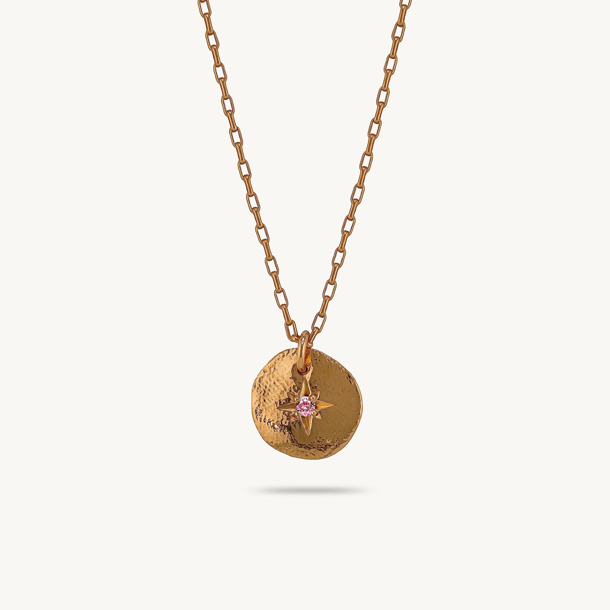A gold-toned chain necklace with a textured circular pendant featuring a small pink starburst charm, shown against a light cream background.