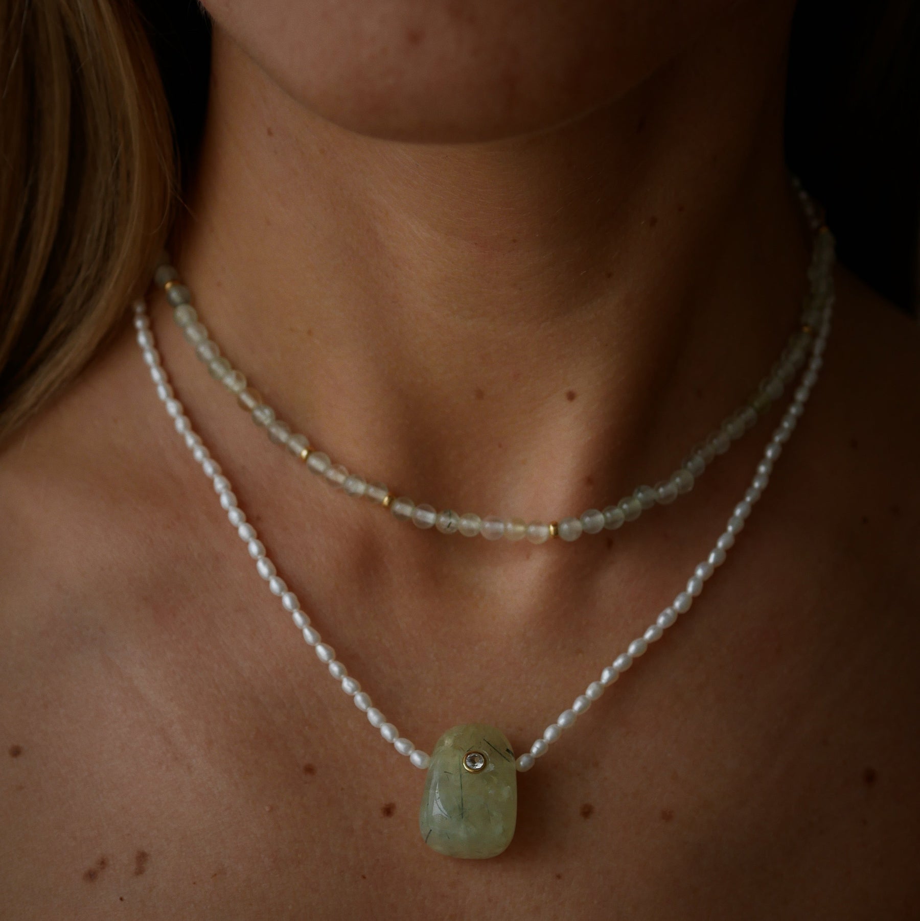 A close-up of a person's neck and upper chest wearing two necklaces. Pearl necklace with a light green rectangular pendant and a delicate prehnite beaded strand layered above it.