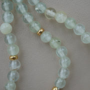 A close-up of a beaded strand featuring pale greenish-white prehnite beads separated by small gold-plated silver spacers, laid on a neutral light beige textured surface.