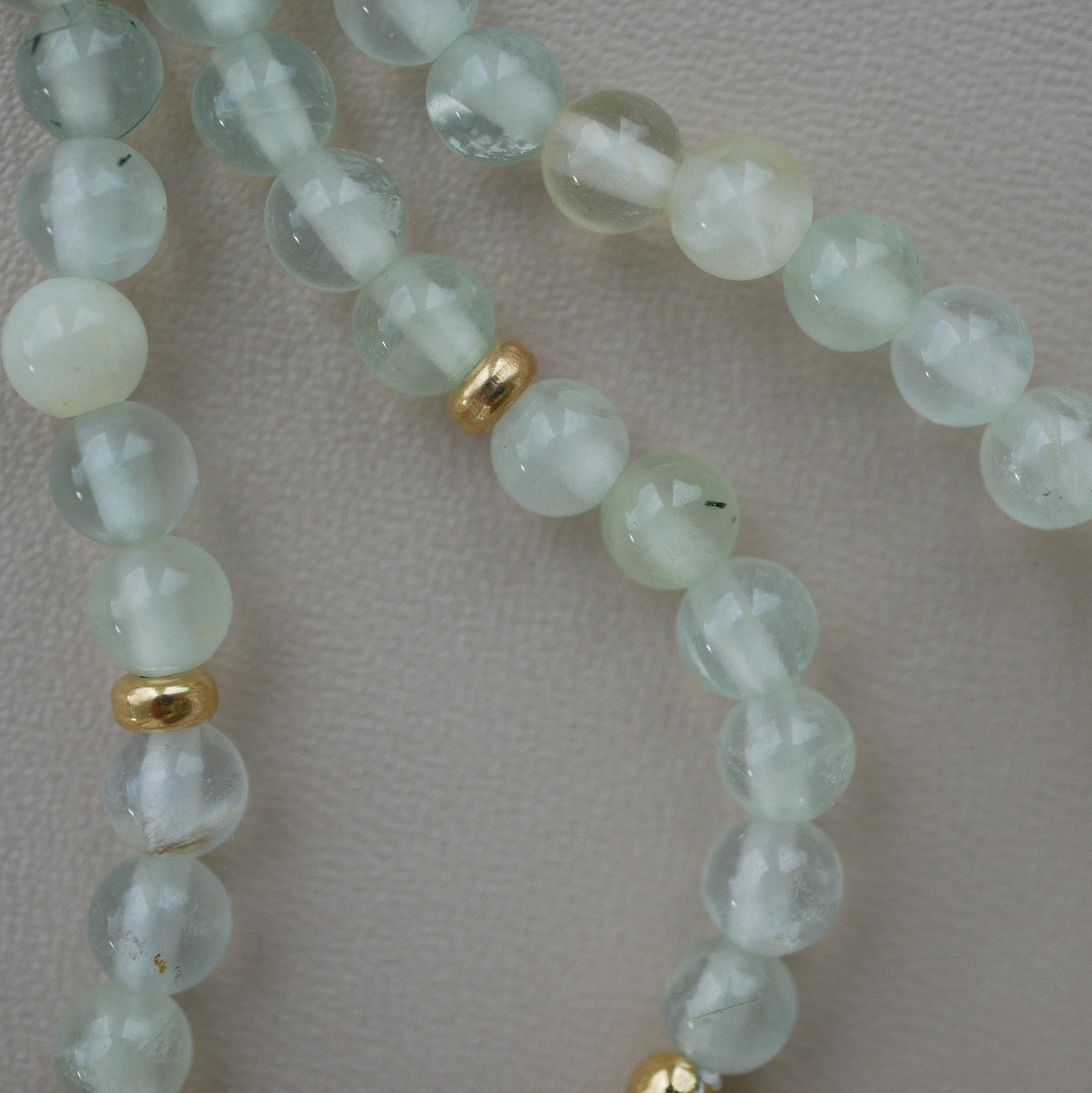 A close-up of a beaded strand featuring pale greenish-white prehnite beads separated by small gold-plated silver spacers, laid on a neutral light beige textured surface.