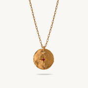 A gold-plated silver pendant with an uneven, organic texture. At its center sits a red sapphire, set within a crisp starburst etching.
