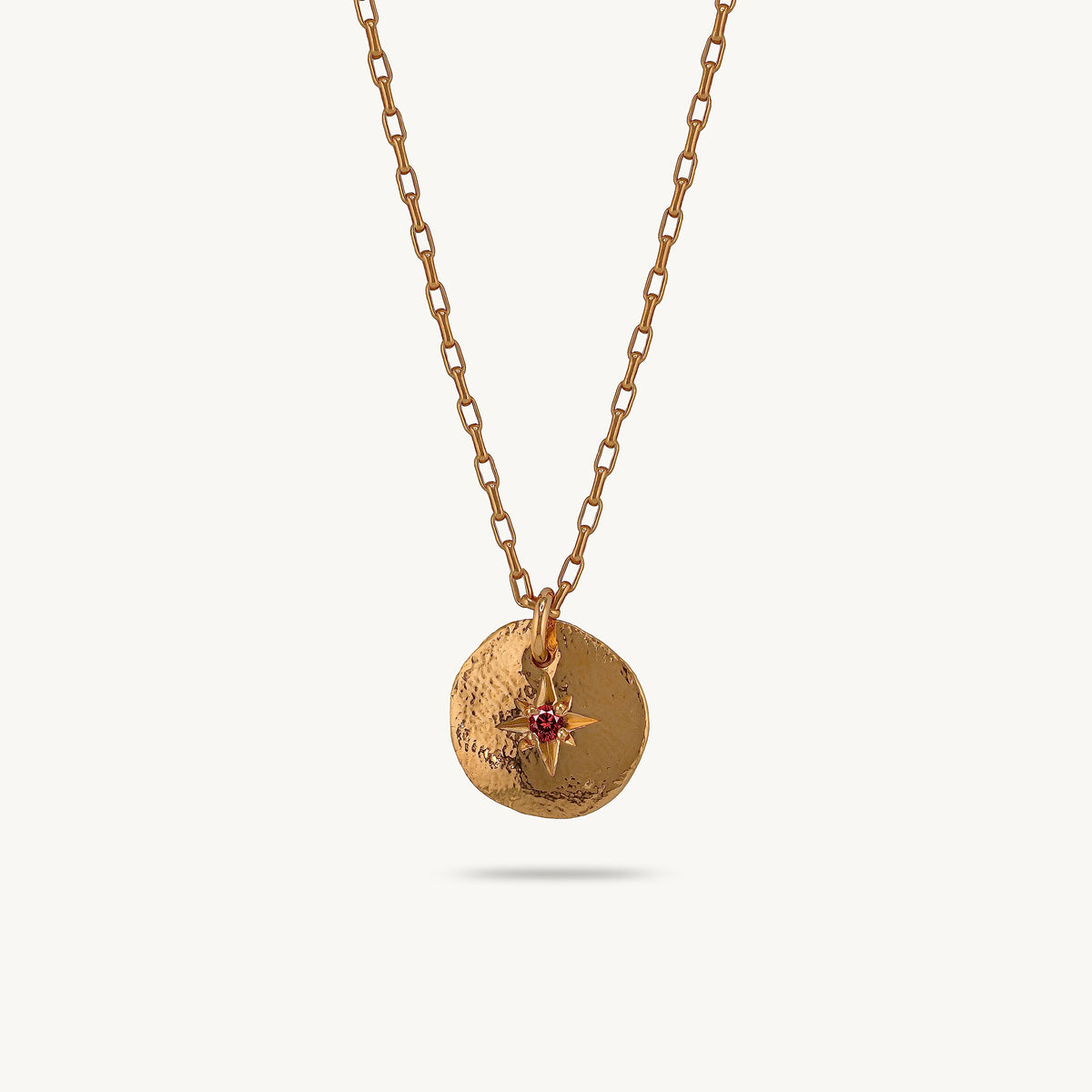 A gold-toned chain necklace with a textured circular pendant featuring a small red sapphire, shown against a light cream background.