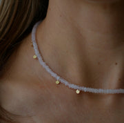 A close-up of a delicate beaded necklace worn on a person's neck. The necklace features pale pinkish-white faceted beads with small gold disc charms and a tiny silver-toned spacer.