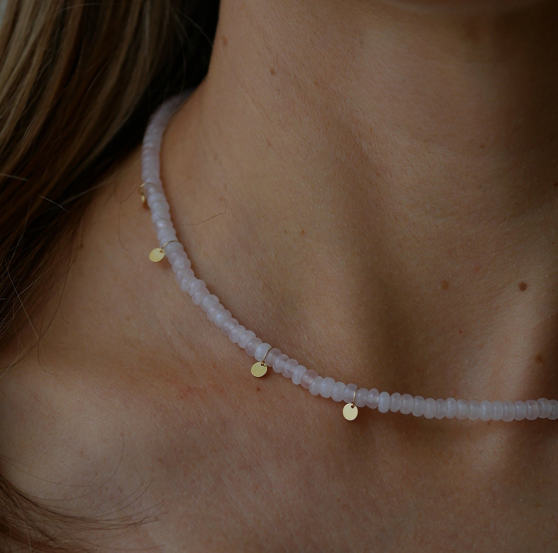 A close-up of a delicate beaded necklace worn on a person's neck. The necklace features pale pinkish-white faceted beads with small gold disc charms and a tiny silver-toned spacer.