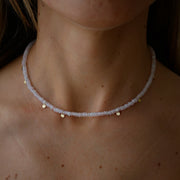A close-up of a person's neck and upper chest wearing a delicate beaded necklace. The necklace consists of small pale pinkish-lilac quartz beads with small gold disc charms spaced along the front.