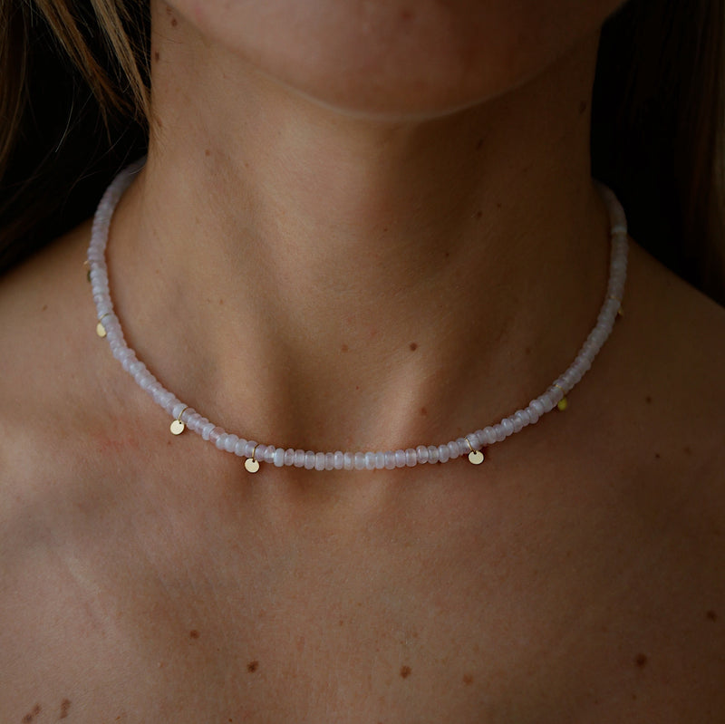A close-up of a person's neck and upper chest wearing a delicate beaded necklace. The necklace consists of small pale pinkish-lilac quartz beads with small gold disc charms spaced along the front.