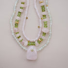 A multi beaded necklaces featuring light pink and white beads, green gemstone beads, and small gold spacer accents, with a large light pink stone pendant at the center, displayed on a soft pink background.