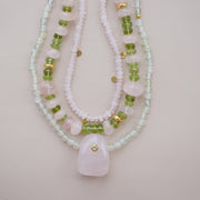 A multi beaded necklaces featuring light pink and white beads, green gemstone beads, and small gold spacer accents, with a large light pink stone pendant at the center, displayed on a soft pink background.