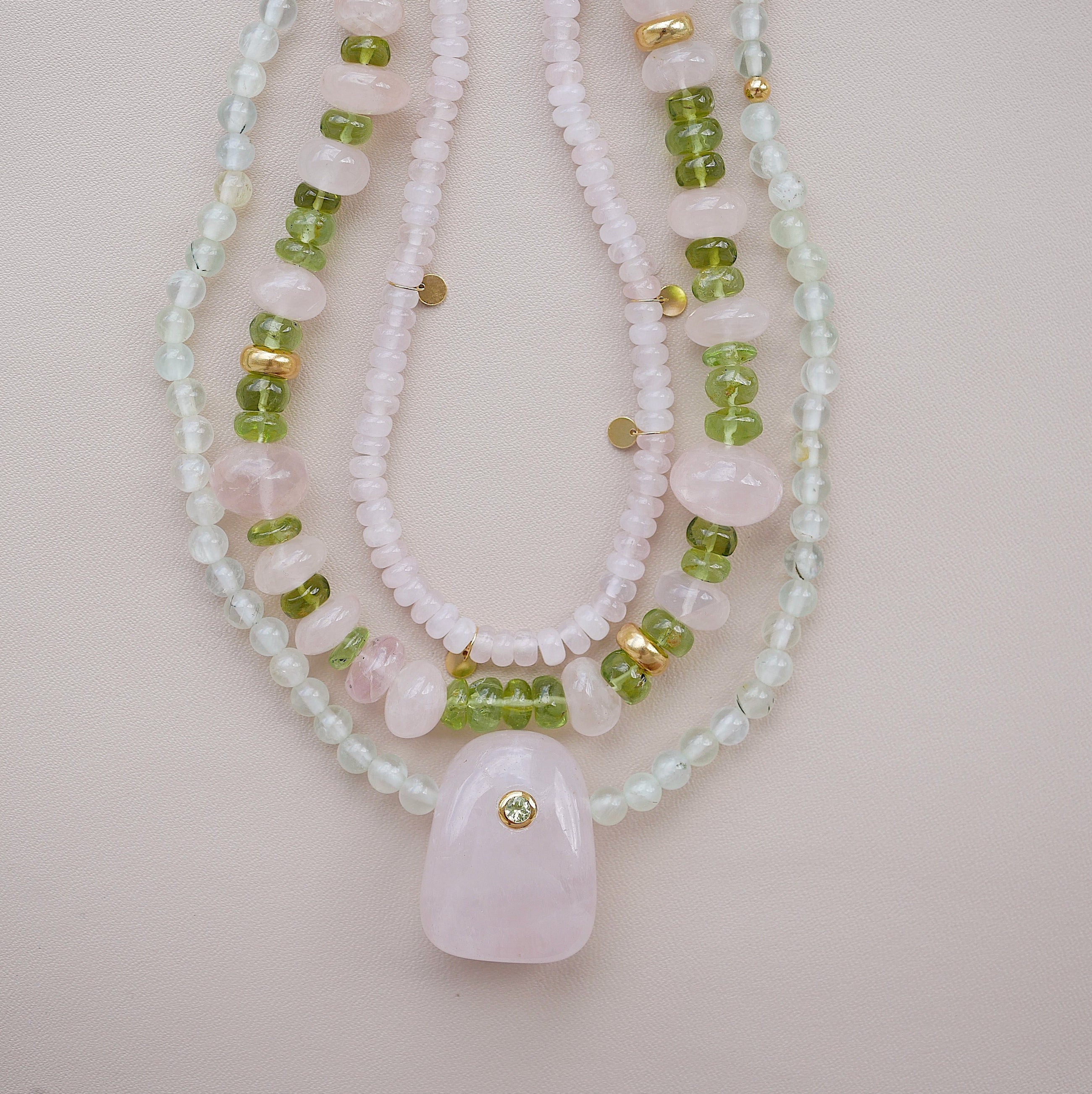A multi beaded necklaces featuring light pink and white beads, green gemstone beads, and small gold spacer accents, with a large light pink stone pendant at the center, displayed on a soft pink background.