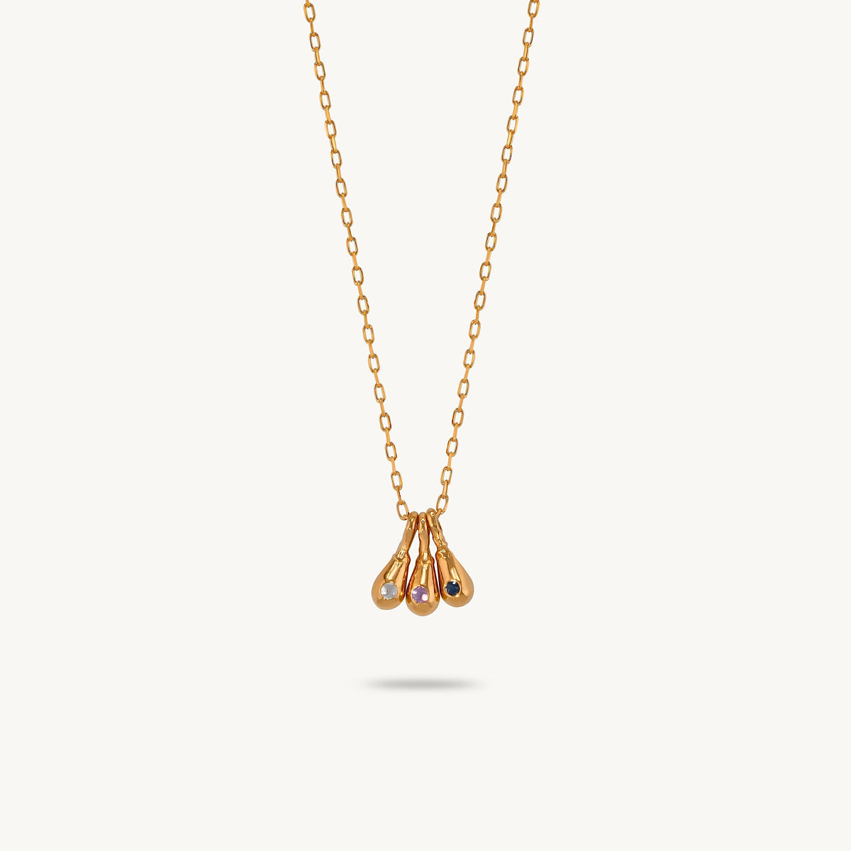 A delicate gold-plated silver necklace with a pendant featuring three teardrop-shaped gold drops, one of which is set with a small iridescent sapphire stone, displayed against a light cream background.