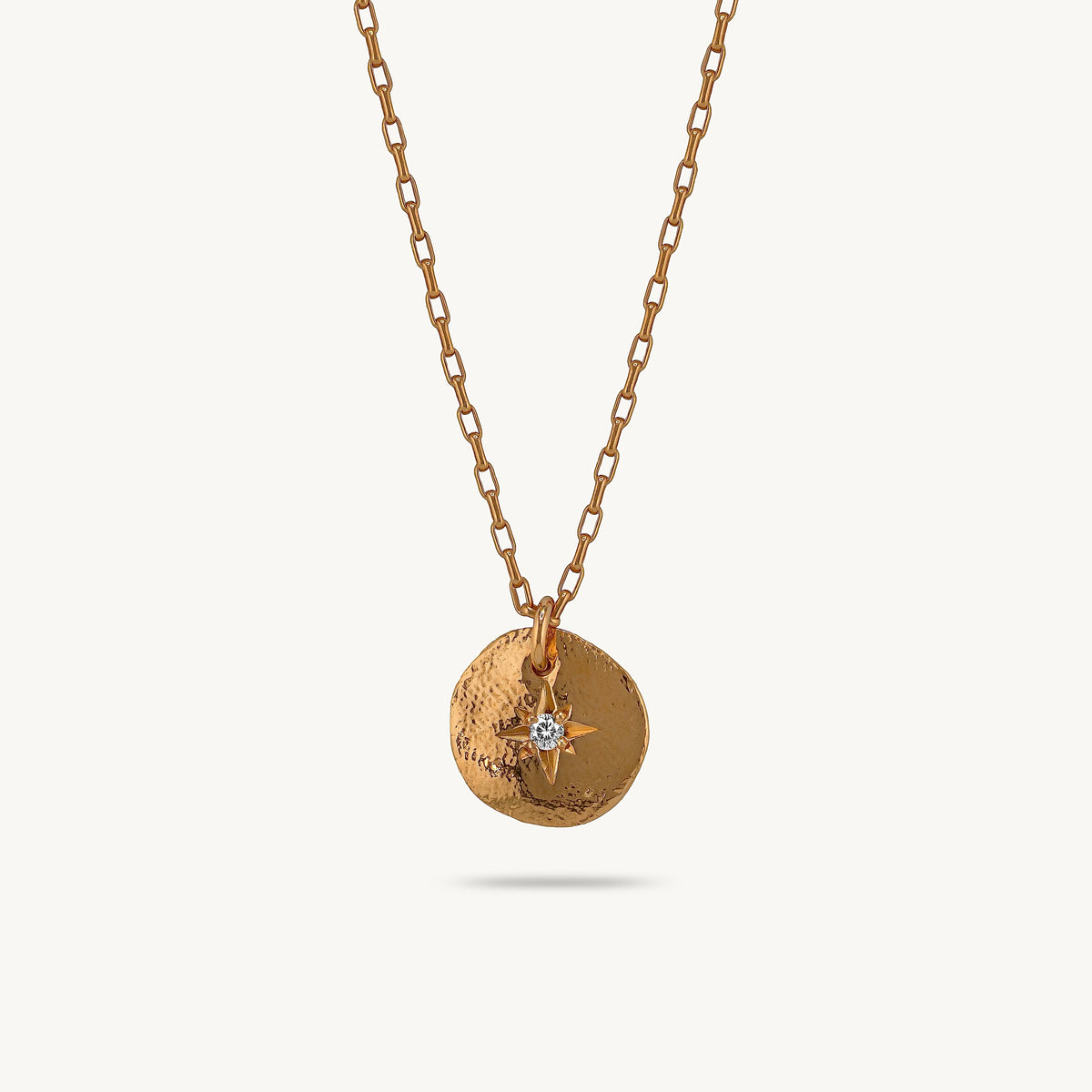 A gold chain necklace featuring a textured circular pendant with a small gold compass rose and a clear central stone, shown against a light cream background.