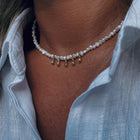 A close-up of a person wearing a delicate pearl necklace with small gold teardrop charms, styled with a light blue linen shirt.