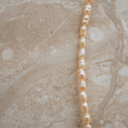 A close-up of a strand of irregular, baroque-style freshwater pearls in a warm champagne-gold color, laid diagonally across a beige marble surface.