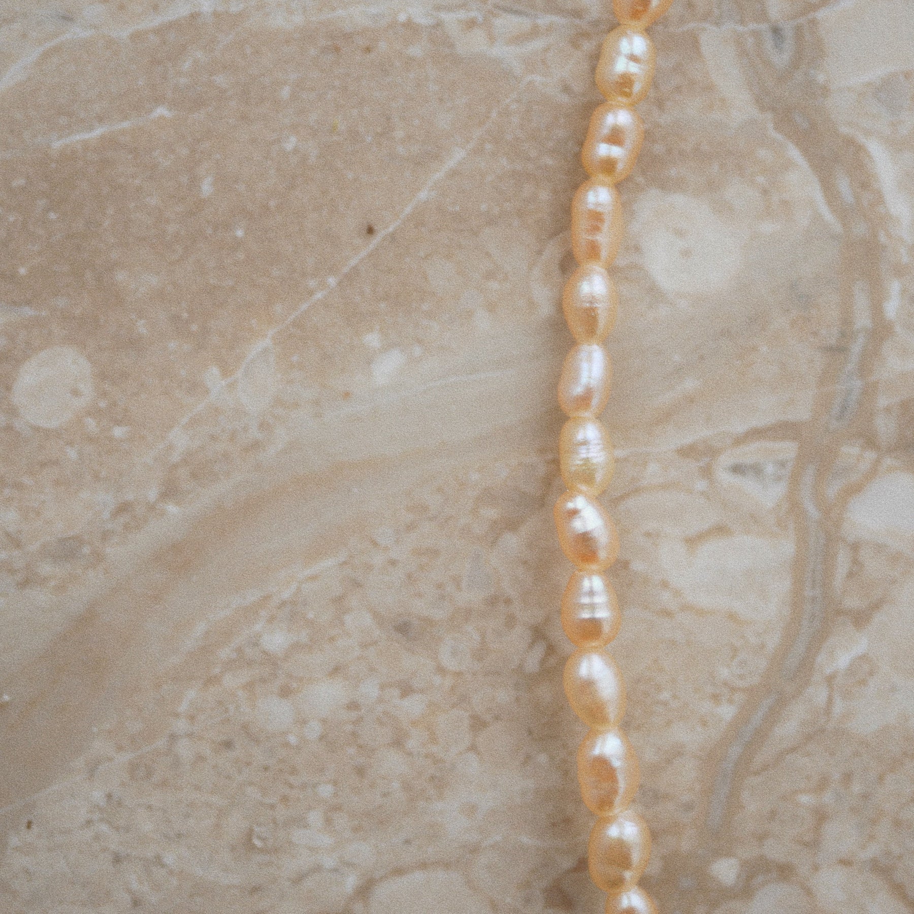 A close-up of a strand of irregular, baroque-style freshwater pearls in a warm champagne-gold color, laid diagonally across a beige marble surface.
