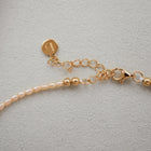 Close-up of a delicate necklace with small pale pink pearls on a gold-plated silver chain, featuring a lobster clasp, extension chain, and a round engraved gold tag, set against a textured light gray background.
