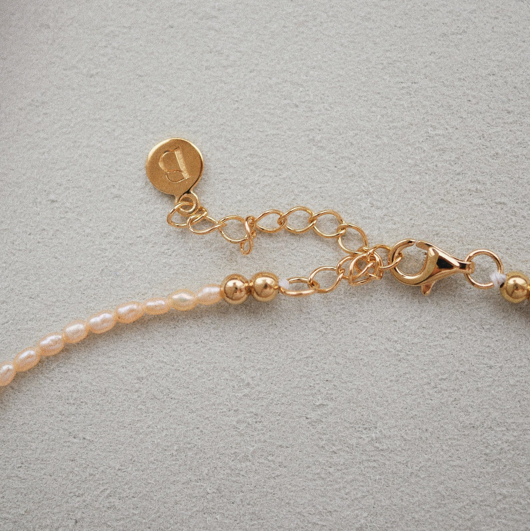 Close-up of a delicate necklace with small pale pink pearls on a gold-plated silver chain, featuring a lobster clasp, extension chain, and a round engraved gold tag, set against a textured light gray background.