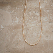 A delicate strand necklace made of small, round, cream-colored pearls, laid out in a U-shape on a beige marble surface.