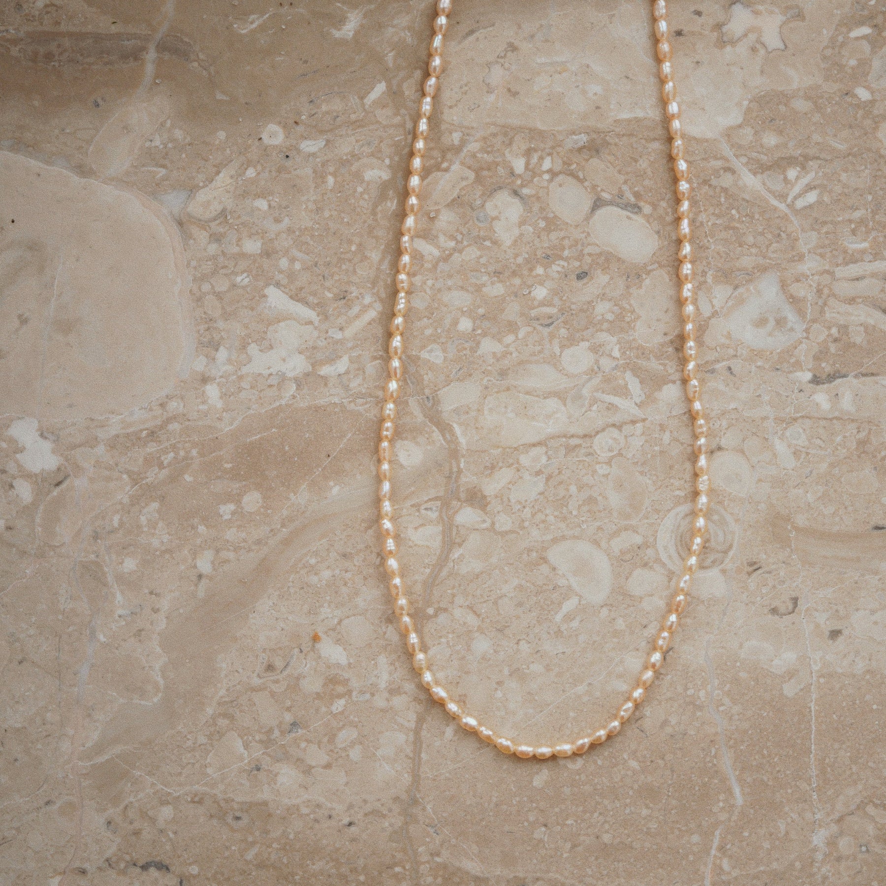 A delicate strand necklace made of small, round, cream-colored pearls, laid out in a U-shape on a beige marble surface.