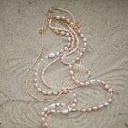 Pearl necklaces with pink, white, and cream pearls, accented with gold-tone chain and clasp, laid out on a sandy surface.