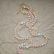 Pearl necklaces with pink, white, and cream pearls, accented with gold-tone chain and clasp, laid out on a sandy surface.