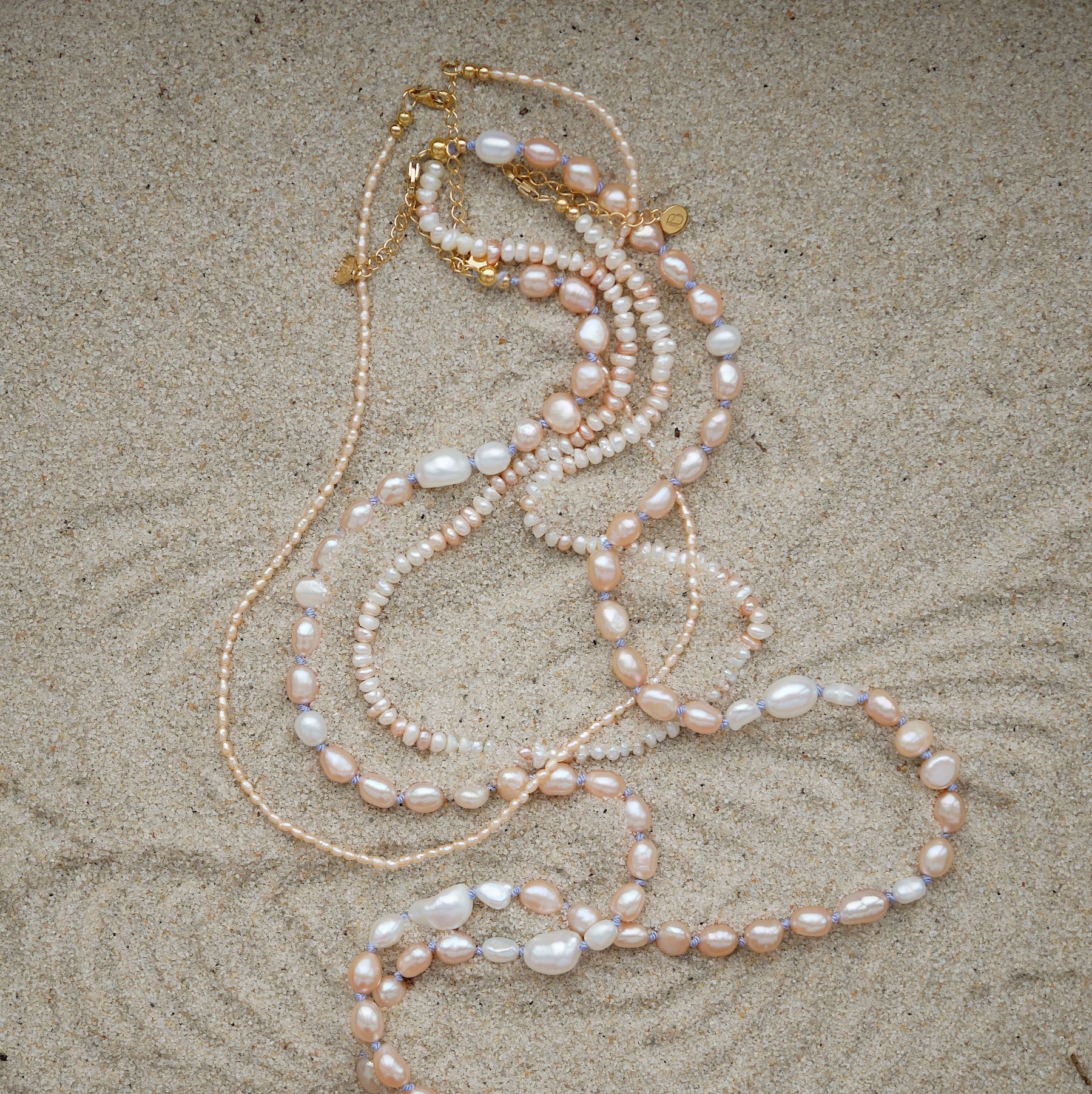 Pearl necklaces with pink, white, and cream pearls, accented with gold-tone chain and clasp, laid out on a sandy surface.