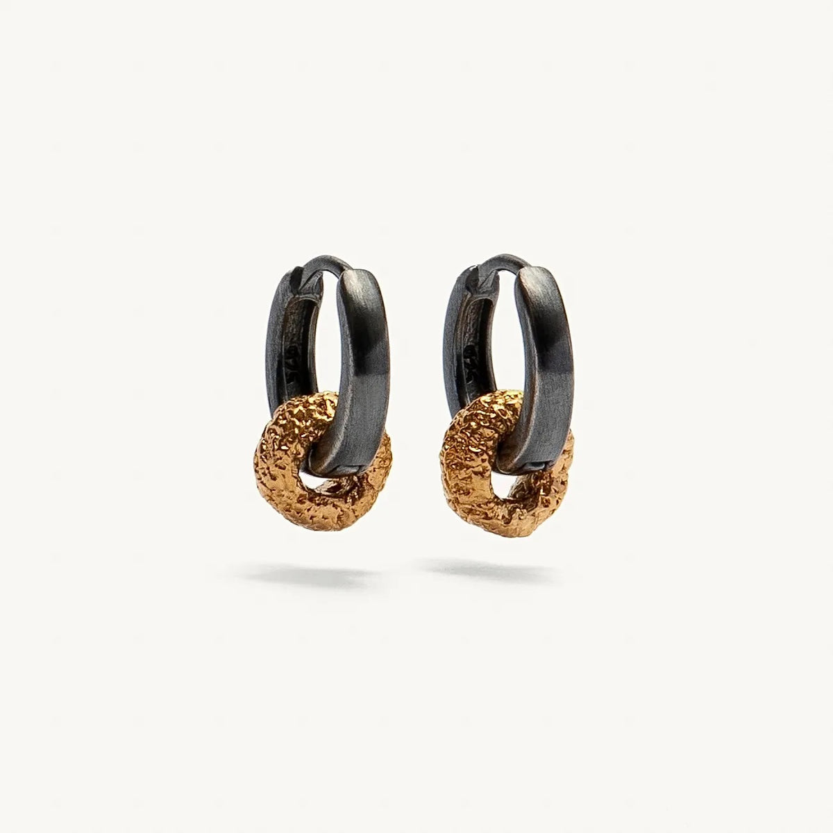 A pair of small huggie hoop earrings with a matte black top half and a textured gold bottom half, shown on a clean off-white background.