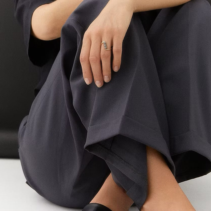 A close-up of a person wearing dark charcoal gray wide-leg trousers, sitting with their legs crossed. The person is wearing a yellow gold ring with three grey faceted diamonds on their finger. The background is a clean, minimalist black and white studio setting.