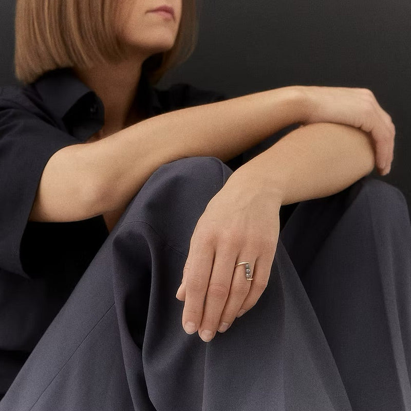 A close-up lifestyle shot of a model wearing a gold & grey diamonds ring with a vertical black inlay, sitting in a dark navy shirt and trousers against a dark gray background.