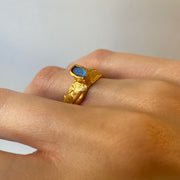A gold-plated bronze ring with a textured band and a blue labradorite gemstone set atop it, worn on a person's finger.