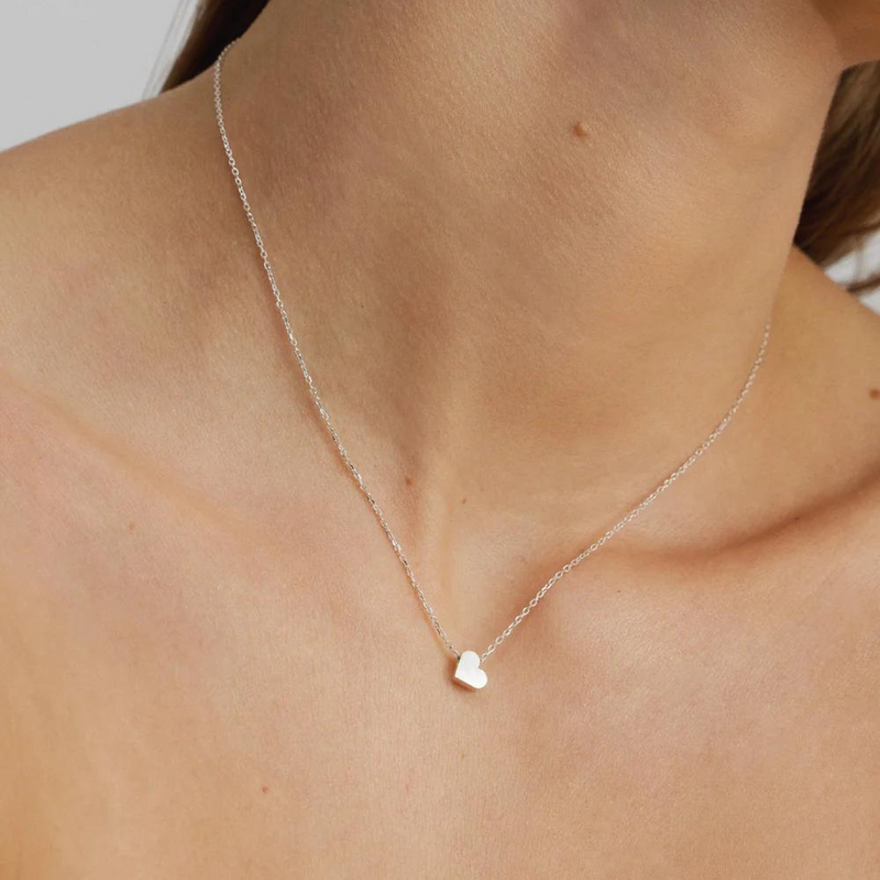 A silver necklace with a flat, silver heart-shaped pendant, displayed on a person's neck.