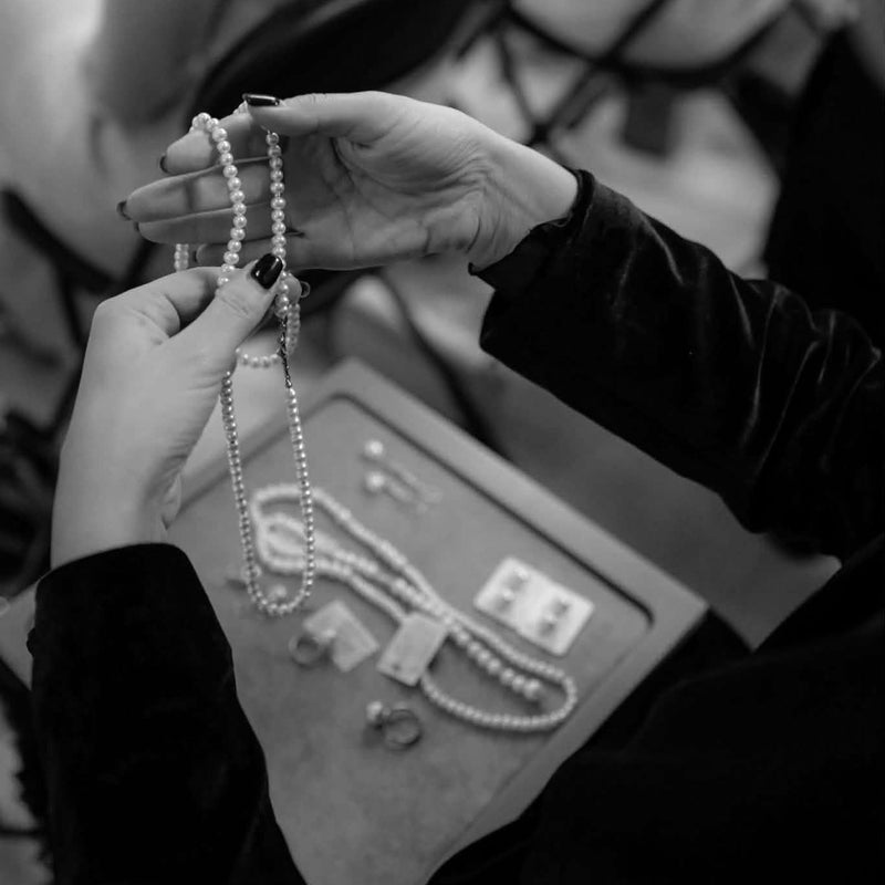 A black and white close-up of a person holding a pearl necklace over a jewelry display tray, with other jewelry and blurred people in the background.