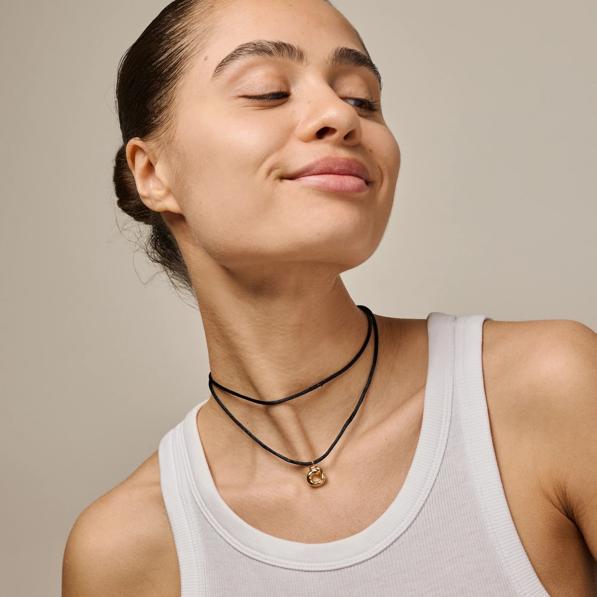 Woman wearing a white tank top and black necklaces with a gold pendant on a beige background