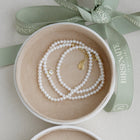 A delicate pearl necklace with gold-toned hardware and a small clear topaz charm, presented inside an open round jewelry box with a sage green Birsenate ribbon.