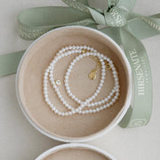 A delicate pearl necklace with gold-toned hardware and a small clear topaz charm, presented inside an open round jewelry box with a sage green Birsenate ribbon.