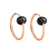 A pair of rose gold hoop earrings with a small, faceted dark grey diamond bead attached near the top right of each hoop, shown against a plain white background.