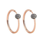 A pair of rose gold hoop earrings with a small, faceted dark grey diamond bead attached near the top right of each hoop, shown against a plain white background.