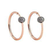 A pair of rose gold hoop earrings with a small, faceted dark grey diamond bead attached near the top right of each hoop, shown against a plain white background.